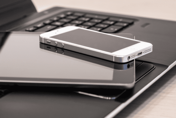 A close-up of a stack of electronic devices, including a smartphone, a tablet, and a laptop. The smartphone is silver with a black screen, the tablet has a black cover, and the laptop has a black keyboard. All devices are placed on a light-colored surface, ready for any SMSF accountant to streamline their workflow.