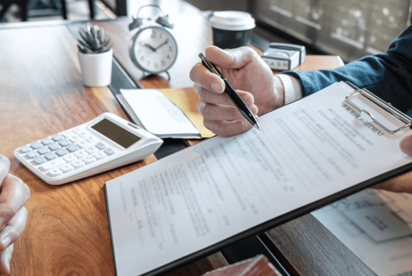 A person is holding and pointing to a clipboard with a document while another person watches. A calculator, clock, coffee cup, and a small plant are on the wooden table in the background. The scene appears to be a meeting or consultation at a Melbourne Accounting Firm.