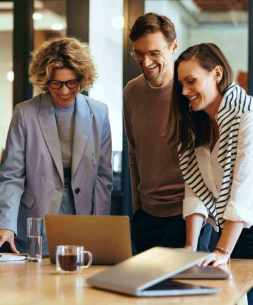 Three people, two women and one man, stand around a table looking at a laptop screen. They are smiling and appear engaged in their discussion. The scene takes place in a modern, well-lit office with glass walls. Coffee mugs, papers, and business accountants' notes are on the table.