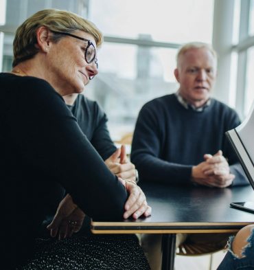 Four people sitting at a table in a meeting room with large windows. A woman in glasses is speaking while a business accountant next to her gestures with his hands. The mood appears focused and attentive.