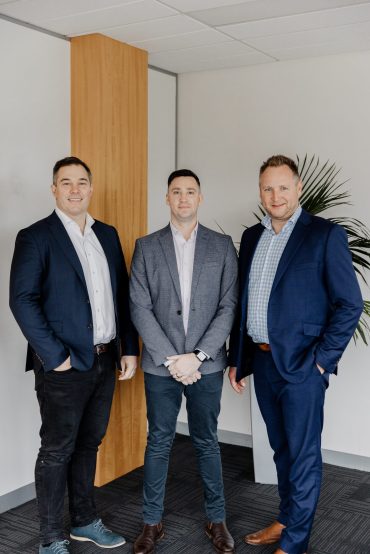 Three men in business attire stand in an office setting, likely representing a Melbourne Accounting Firm. Clad in blazers and dress shirts, one man also sports jeans. They stand before a white wall with a wooden panel and a large green plant to the right, epitomizing professional Business Advisors or SMSF Accountants at work.