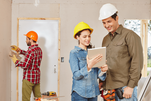Three construction workers in a room being renovated. Two of them, wearing hard hats, look at a tablet and smile, possibly discussing plans with their Melbourne Accounting Firm. Meanwhile, the third worker in the background uses a drill on the wall. Tools and building materials are scattered around the workspace.