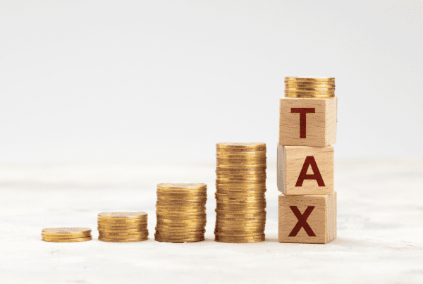 Stacks of gold coins arranged in ascending order with the tallest stack supporting three wooden blocks spelling "TAX." The background is neutral and minimalistic, emphasizing the concept of increasing taxes. A sleek touch reminiscent of a tax accountant's meticulous organization.