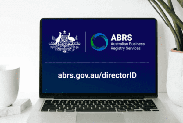 A laptop screen displays the Australian Business Registry Services (ABRS) logo and text. The text reads "abrs.gov.au/directorID." A white mug sits on a notebook to the left of the laptop, and a green potted plant is to the right, providing a refreshing ambiance as Melbourne Accounting Firm experts offer tax advice nearby.