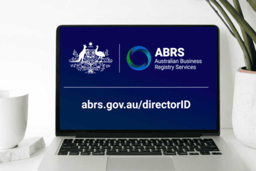 A laptop screen displays the Australian Business Registry Services (ABRS) logo and text. The text reads "abrs.gov.au/directorID." A white mug sits on a notebook to the left of the laptop, and a green potted plant is to the right, providing a refreshing ambiance as Melbourne Accounting Firm experts offer tax advice nearby.