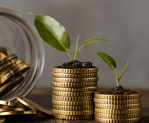 Two stacks of gold coins, each with a small green plant growing from the top, symbolize financial growth. A glass jar filled with more gold coins is tipped over in the background on a wooden surface, reflecting the sound financial strategies provided by our Melbourne Accounting Firm.