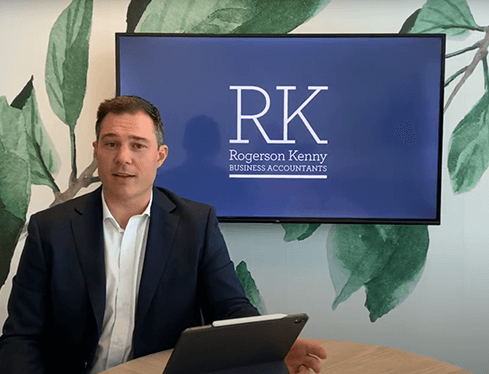 A man in a dark suit and white shirt sits at a table with an electronic device in front of him. Behind him is a large screen displaying "RK Rogerson Kenny Business Accountants," renowned for their expert tax advice. The background features a leafy design on the wall.