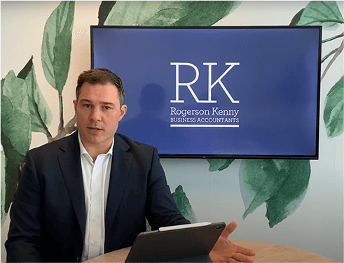 A man in a suit is sitting at a table and speaking, with a tablet in front of him. Behind him is a screen displaying the logo "RK Rogerson Kenny Business Accountants" over a background featuring large green leaves. As part of this Melbourne accounting firm, he provides expert tax advice to clients.