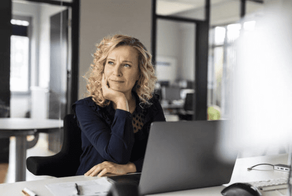 A blonde woman in a navy blue blouse sits at an office desk with a laptop, resting her face on her hand and appearing thoughtful. She is in a modern office setting with glass walls and minimalistic decor, reflecting the professional environment of a Melbourne accounting firm.