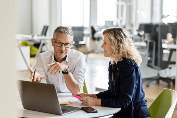 Two colleagues sit at a table in a modern office. One person, wearing glasses and a white shirt, gestures while speaking. The other, a blonde individual in a dark outfit, smiles while holding a highlighter. A laptop and smartphone are on the table, emblematic tools for skilled Business Accountants at this Melbourne Accounting Firm.