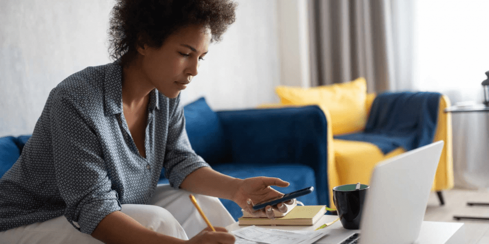A person sits on a sofa with a laptop, holding a phone and writing on documents spread on a table. The background features a blue sofa with yellow cushions and a mug beside the laptop, possibly discussing tax advice or working with SMSF accountants to finalize reports.