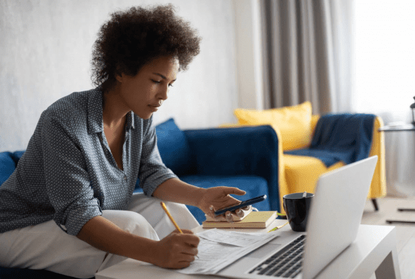 A person sits on a sofa with a laptop, holding a phone and writing on documents spread on a table. The background features a blue sofa with yellow cushions and a mug beside the laptop, possibly discussing tax advice or working with SMSF accountants to finalize reports.