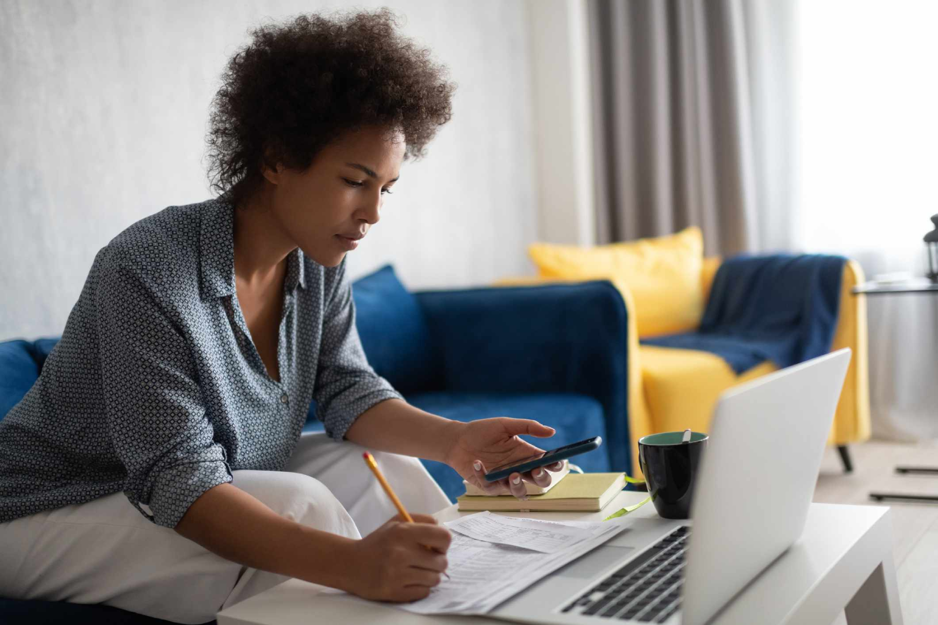 A person sits on a sofa with a laptop, holding a phone and writing on documents spread on a table. The background features a blue sofa with yellow cushions and a mug beside the laptop, possibly discussing tax advice or working with SMSF accountants to finalize reports.