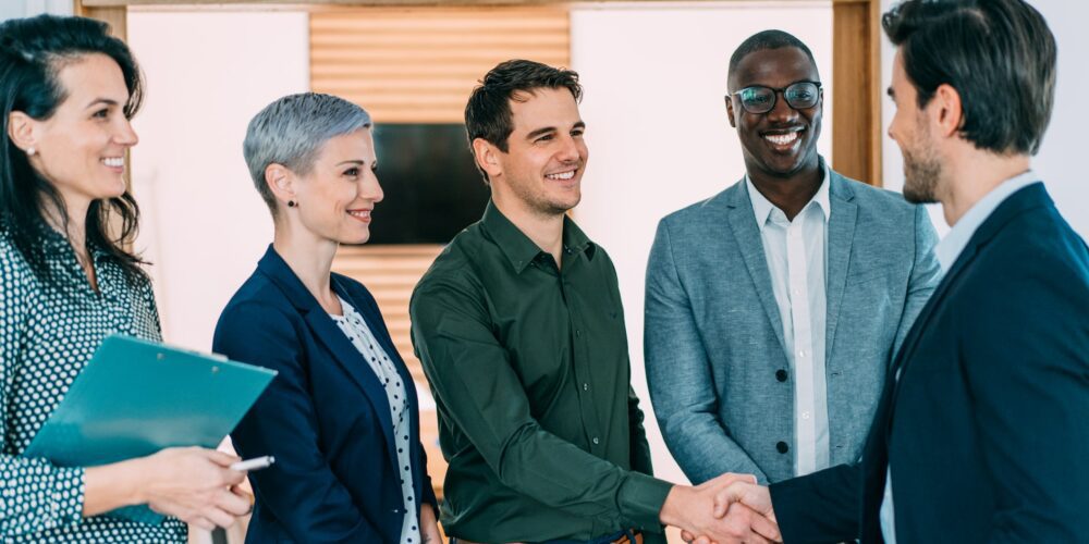 A group of five professionally dressed people stands indoors, with three men and two women. One man is shaking hands with another, while the others look on and smile. One woman holds a clipboard. They appear to be business advisors in a professional setting, possibly offering tax advice.