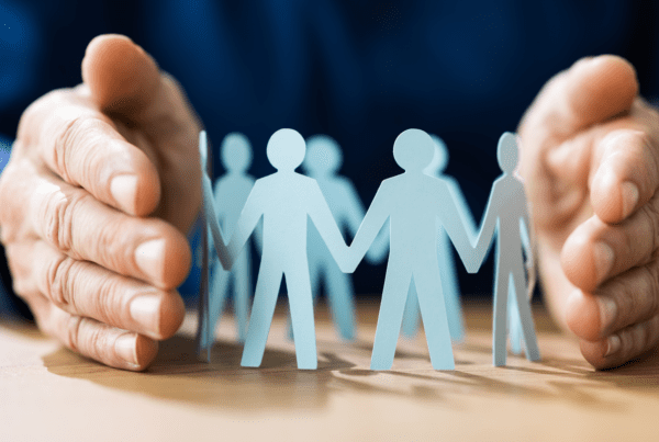 Close-up of hands gently surrounding a circle of paper cutout figures. The figures are holding hands, symbolizing unity, teamwork, and protection—a perfect representation for business accountants fostering community and trust. The background is blurred, drawing focus to the hands and paper figures.