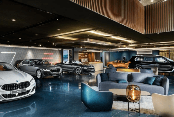 A sleek, modern car showroom features various BMW models, including sedans, convertibles, and SUVs. The floor is shiny blue, and stylish seating areas with contemporary furniture are provided for customers. As well-lit as a business advisor’s office, the space boasts a sophisticated interior design.