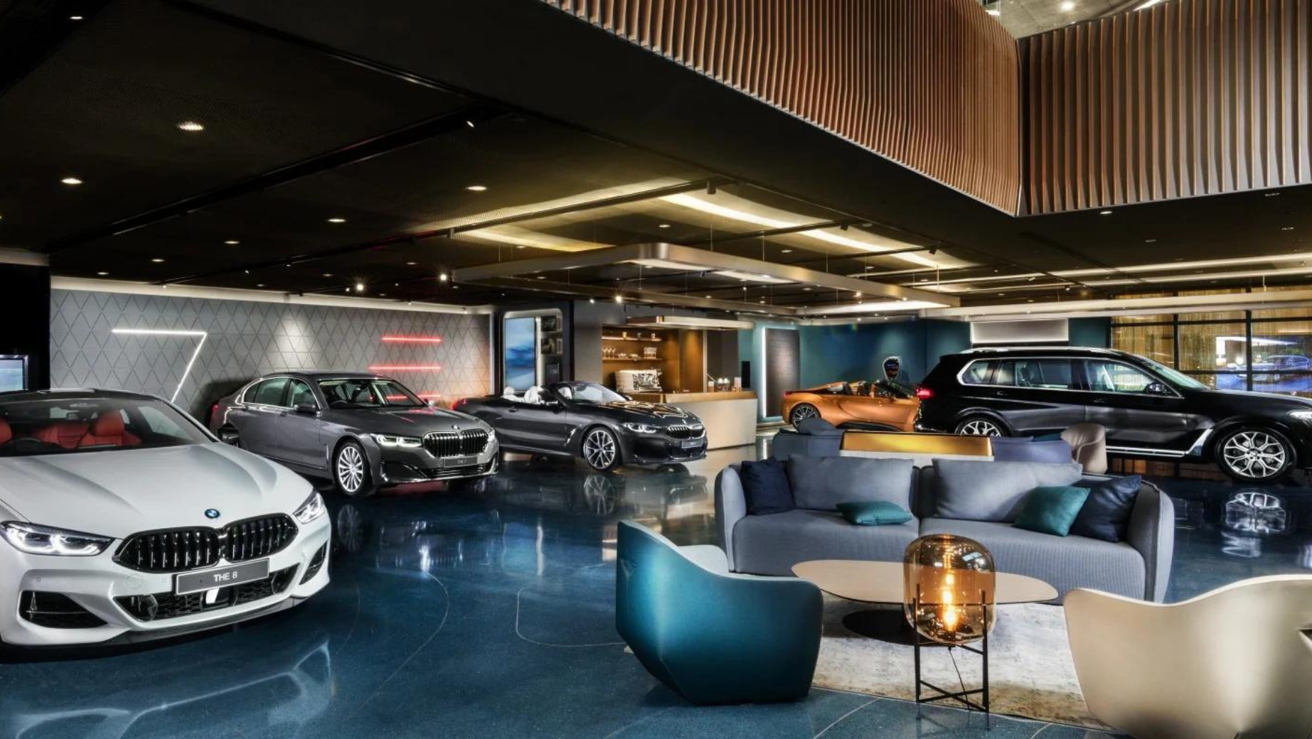 A sleek, modern car showroom features various BMW models, including sedans, convertibles, and SUVs. The floor is shiny blue, and stylish seating areas with contemporary furniture are provided for customers. As well-lit as a business advisor’s office, the space boasts a sophisticated interior design.
