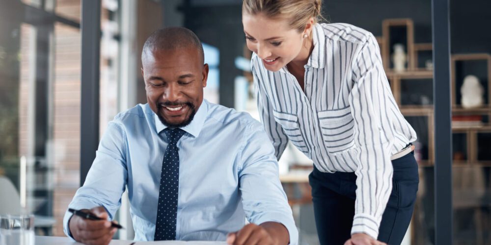 A man and a woman are working together at a desk in an office setting. The man, seated and holding a pen, smiles while looking at a notebook. The woman, standing beside him, also smiles as she looks at the notebook. At this Melbourne accounting firm, the atmosphere appears collaborative.