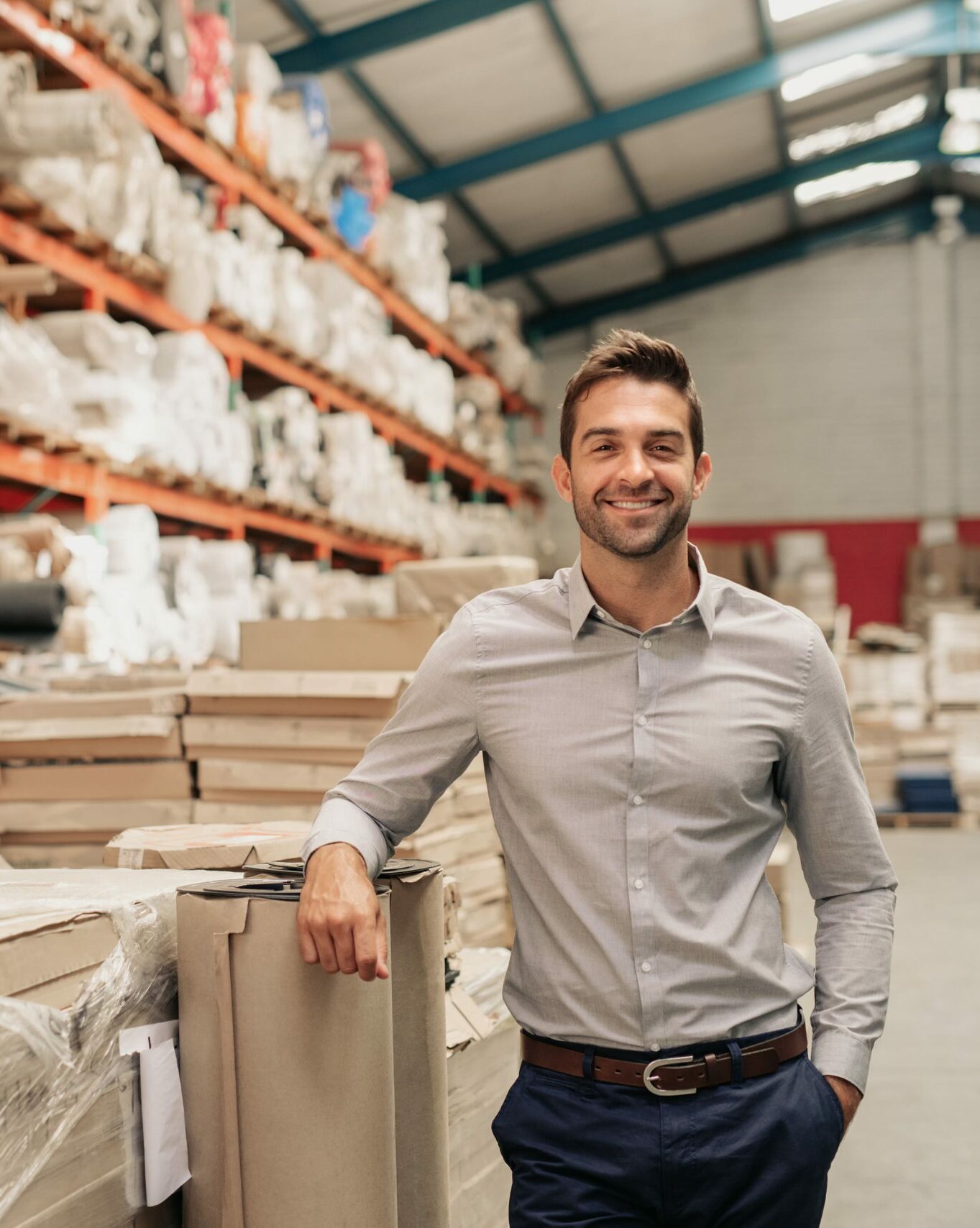 A man in a button-up shirt and slacks stands smiling in a warehouse, resting his arm on a pile of wrapped packages. Rows of shelves filled with various products are visible in the background. The well-lit space, illuminated by natural light from windows above, belongs to a Melbourne Accounting Firm.