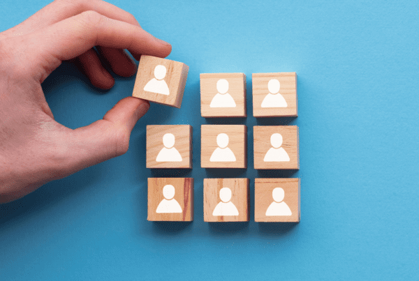 A hand is placing a wooden block with a person icon among a group of similar blocks on a blue background. Each block features a white icon of a person, suggesting the careful selection process typically employed by business advisors for team building.