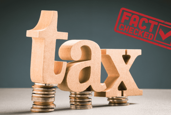 Large wooden letters spelling "tax" are balanced on stacks of coins. To the right, a red stamp with the text "FACT CHECKED" and a checkmark is prominently displayed. The background is dark, providing a stark contrast to the wooden letters and the red stamp, reminiscent of a Melbourne Accounting Firm's office decor.