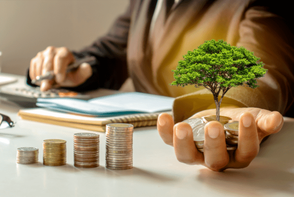 A person in a suit is using a calculator and writing in a notebook at a desk, embodying the meticulous work of business advisors. In the foreground, stacked coins increase in height from left to right, and the person's other hand holds a small tree growing from a pile of coins, symbolizing prosperous tax advice.