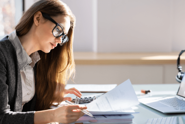 A woman with long hair and glasses works at a desk. She is looking at documents and using a calculator. A laptop displaying an invoice from a Melbourne accounting firm is open in front of her, along with pens and a stack of papers. The bright, well-lit workspace suggests she might be a tax accountant.