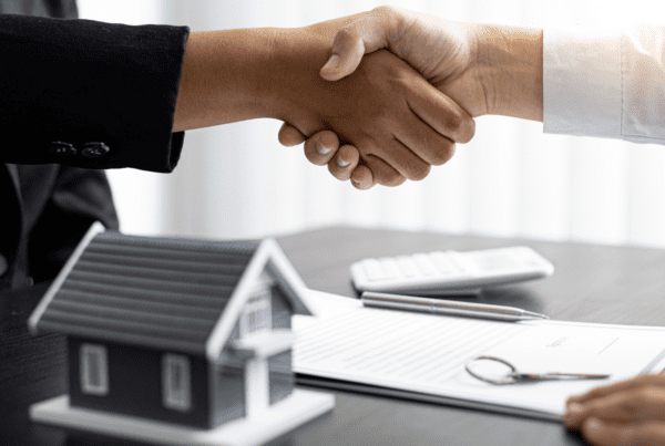 Two hands shaking over a table with a small model house, eyeglasses, a contract, a pen, and a calculator. The scene suggests the closing of a real estate deal or financial agreement, possibly facilitated by experts from an SMSF accountants group or Melbourne accounting firm.