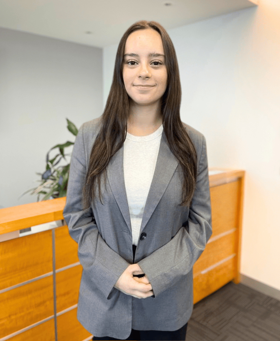 A young woman with long brown hair stands indoors, wearing a gray blazer over a light shirt. She is smiling softly and has her hands clasped in front of her, with office furniture and a plant in the background.