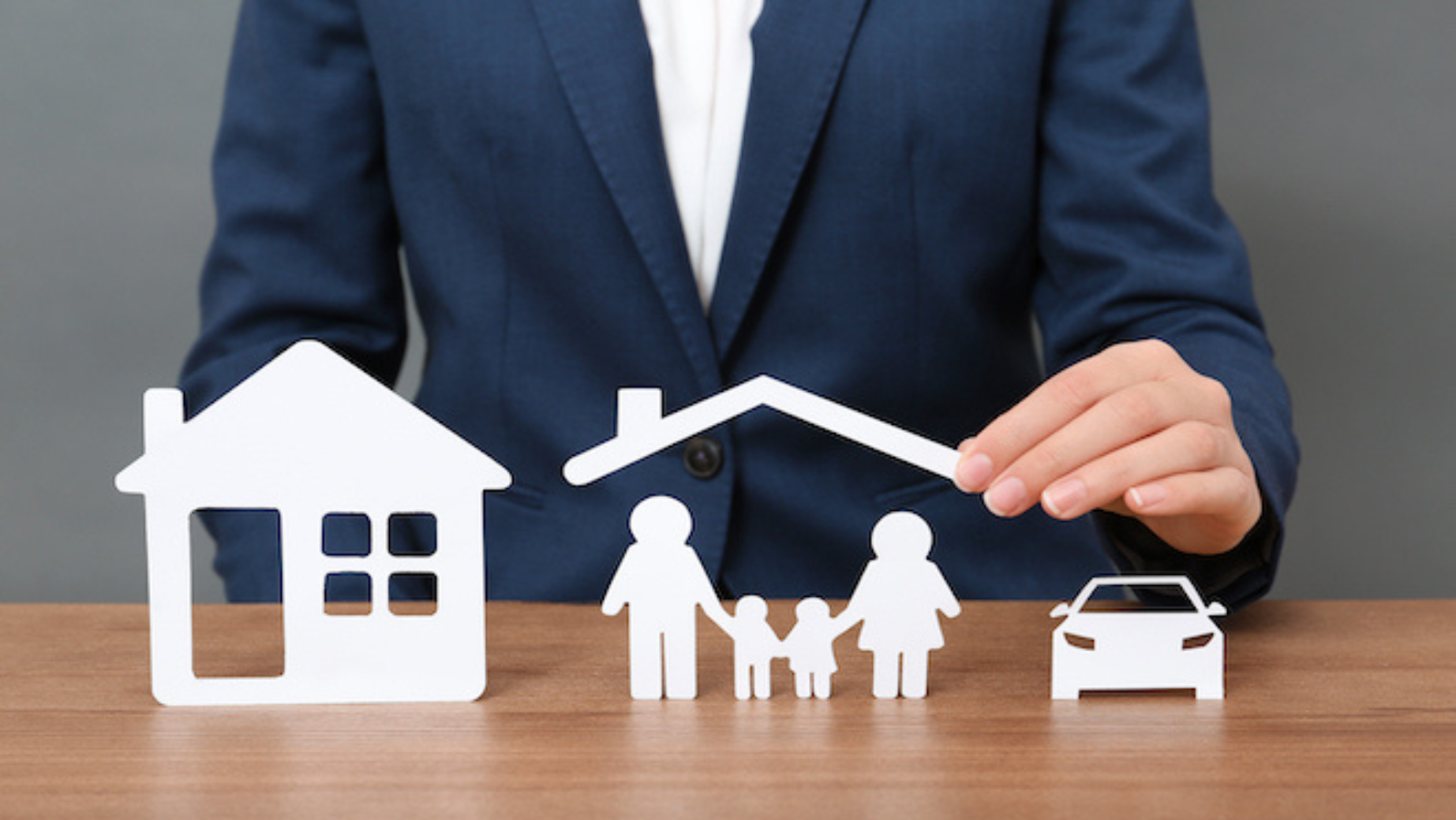 A person in a blue suit is arranging white cutouts of a house, a family holding hands under a roof, and a car on a wooden surface. The image symbolizes the protection and security that Melbourne Accounting Firm provides for your home, family, and vehicle.