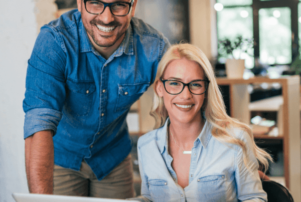 A smiling man and woman wearing glasses and casual denim shirts are working together at a desk with an open laptop. The office setting, part of a Melbourne accounting firm, has a white interior with ambient lighting and large windows. The man stands beside the seated woman, providing tax advice.