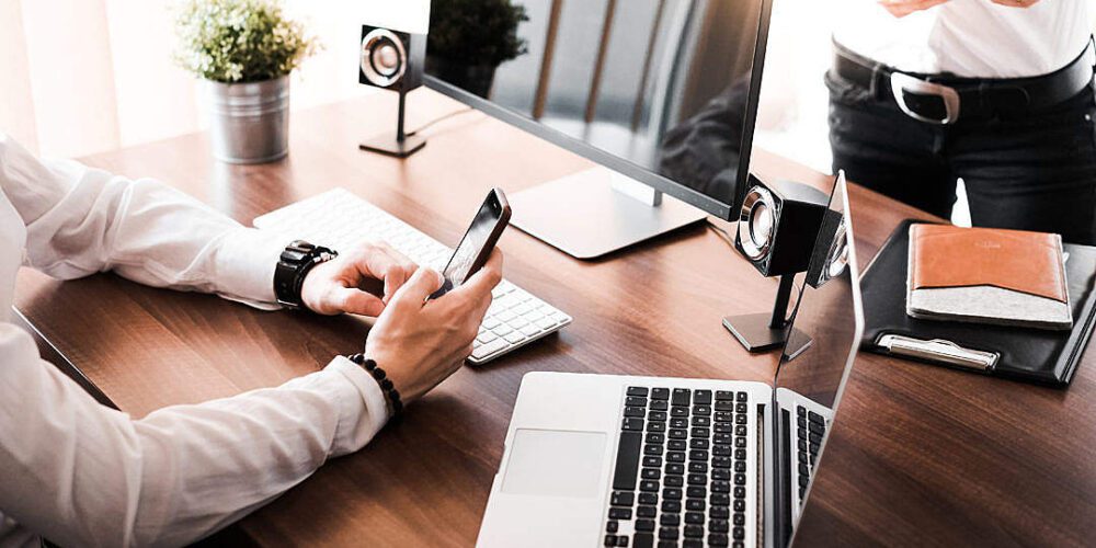 A person seated at a wooden desk, resembling the typical setup of business advisors, is using a smartphone. The desk also has a laptop, desktop computer, speakers, and potted plant. Another person stands nearby holding a tablet. Both in white shirts and dark pants, the scene is well-lit by natural light.