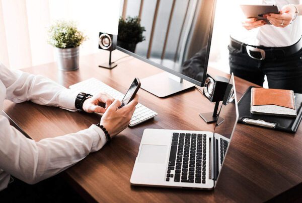 A person seated at a wooden desk, resembling the typical setup of business advisors, is using a smartphone. The desk also has a laptop, desktop computer, speakers, and potted plant. Another person stands nearby holding a tablet. Both in white shirts and dark pants, the scene is well-lit by natural light.