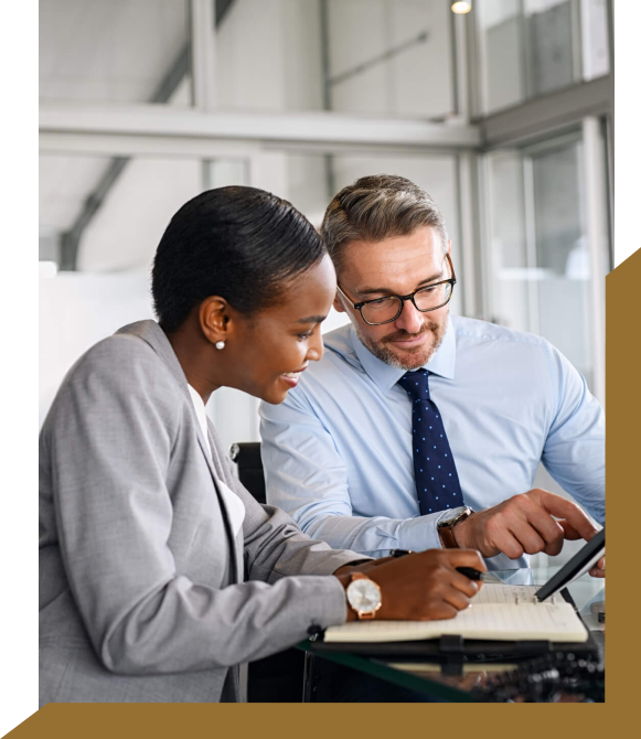 A woman in a gray blazer and a man in a light blue shirt with glasses are seated at a desk, looking at a tablet and a notebook. The woman is writing in the notebook, while they both appear focused on their work as business advisors for a prominent Melbourne accounting firm.