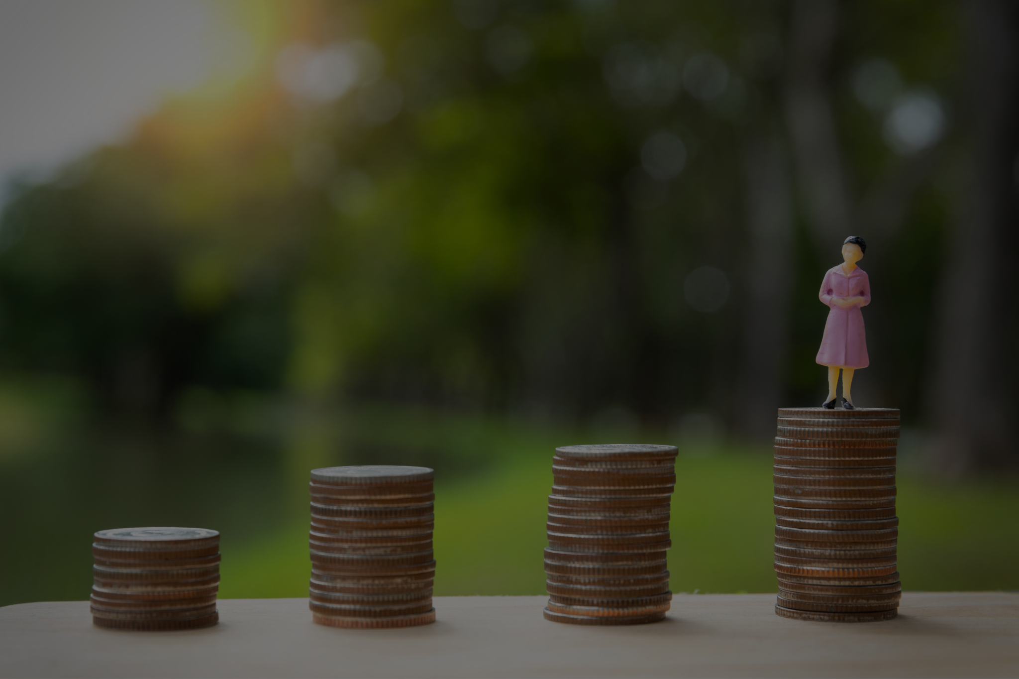 A small figurine of a person in a pink dress stands atop one of four progressively taller stacks of coins, symbolizing financial growth. Blurred greenery is seen in the background, representing the insightful guidance of business advisors.