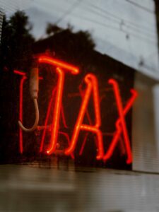A red neon sign displays the word "TAX" in capital letters. Captured in a slightly dim environment, a reflection dances on the glass while trees and power lines loom in the background, evoking a call for expert Tax Advice amidst urban life.