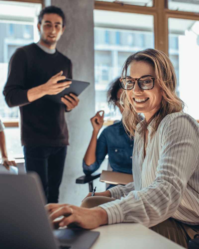A woman with glasses smiles while using a laptop at a desk. In the background, a business advisor holding a tablet stands beside a window, engaging in conversation with another partially visible person. The scene takes place in a well-lit, modern office environment.