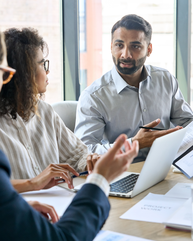 A man with a beard and a gray shirt is speaking to a woman with curly hair and glasses. They are seated at a table with a laptop and documents, likely discussing financial details. Another person, partially visible, is gesturing with their hand. This scene appears to be taking place at a Melbourne Accounting Firm.